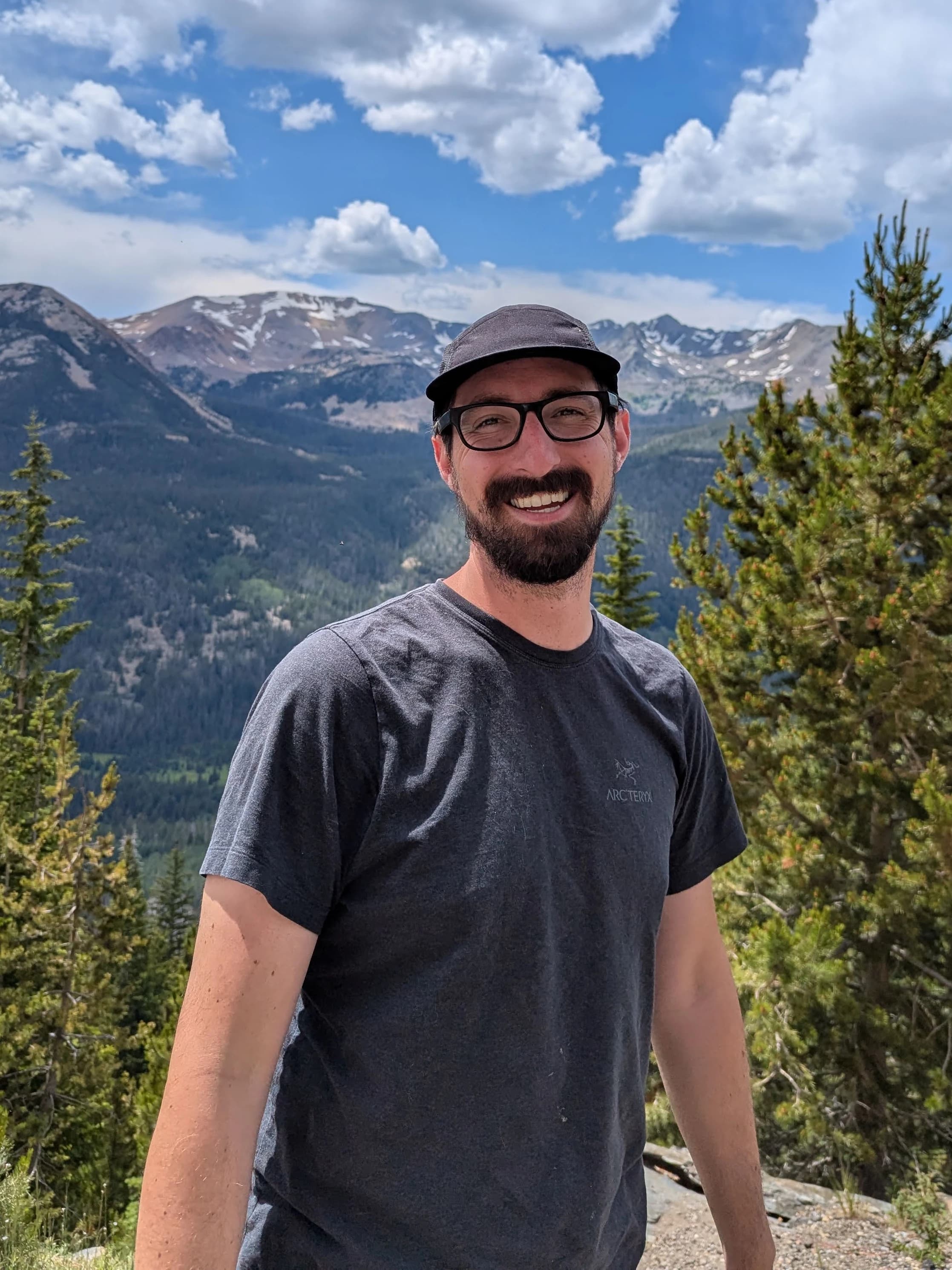 Mike Charpin smiling in a charcoal shirt with a black hat and thick frames black glasses in from of the Rocky Mountains. 2024